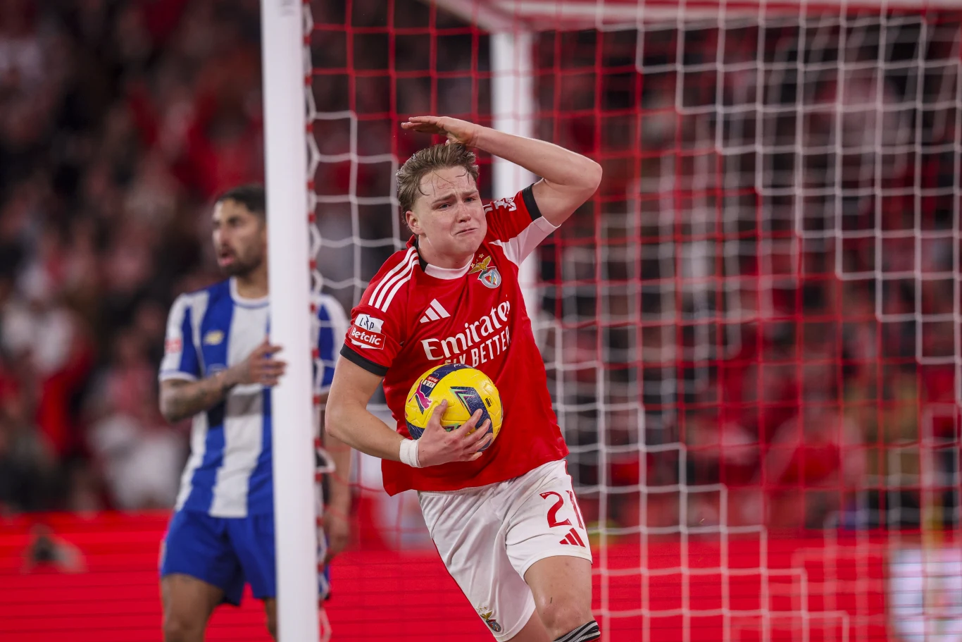 piłkarz w czerwonym stroju drużyny Benfica trzyma piłkę pod pachą i podnosi rękę w geście emocji przy bramce, w tle zawodnik drużyny przeciwnej w niebiesko-białych barwach oraz siatka bramkowa