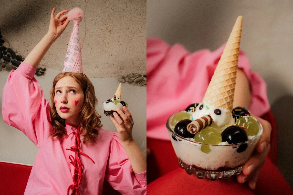 kobieta w różowej bluzce i czapce imprezowej z różowym pomponem trzyma w ręku deser lodowy z waflem, owocami, ciasteczkami i bitą śmietaną ułożonymi dekoracyjnie w szklanej miseczce