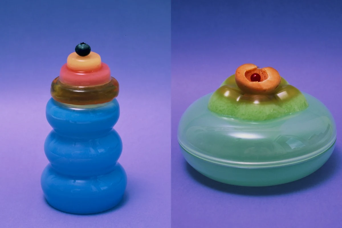 Dwa fantazyjne pojemniki w nasyconych kolorach na fioletowym tle, jeden o niebieskiej, segmentowej formie z owocową dekoracją na wierzchu w postaci borówki i plasterków owoców, drugi o bardziej płaskim, zielonym kształcie z dekoracją z połówki brzoskwi...