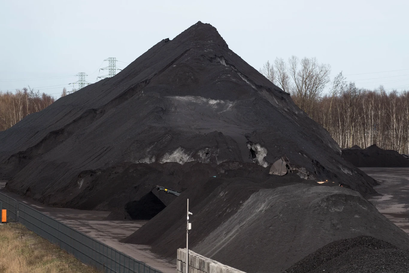 wysoka góra czarnego węgla usypana na otwartym terenie przemysłowym, w tle widoczne bezlistne drzewa oraz słupy energetyczne