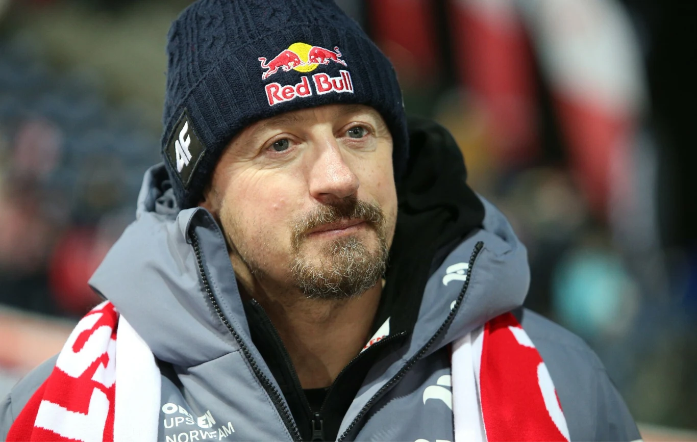 mężczyzna w zimowej kurtce i czapce z logo Red Bull oraz szalikiem kibica na szyi, na tle rozmytych biało-czerwonych flag