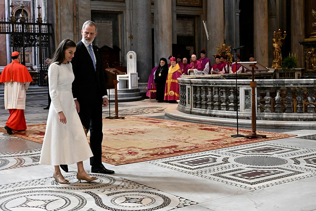 Para ubrana elegancko idzie po ozdobnej posadzce w dużej, bogato zdobionej świątyni, w tle grupa duchownych w purpurowych i złotych szatach gromadzi się przy ołtarzu.
