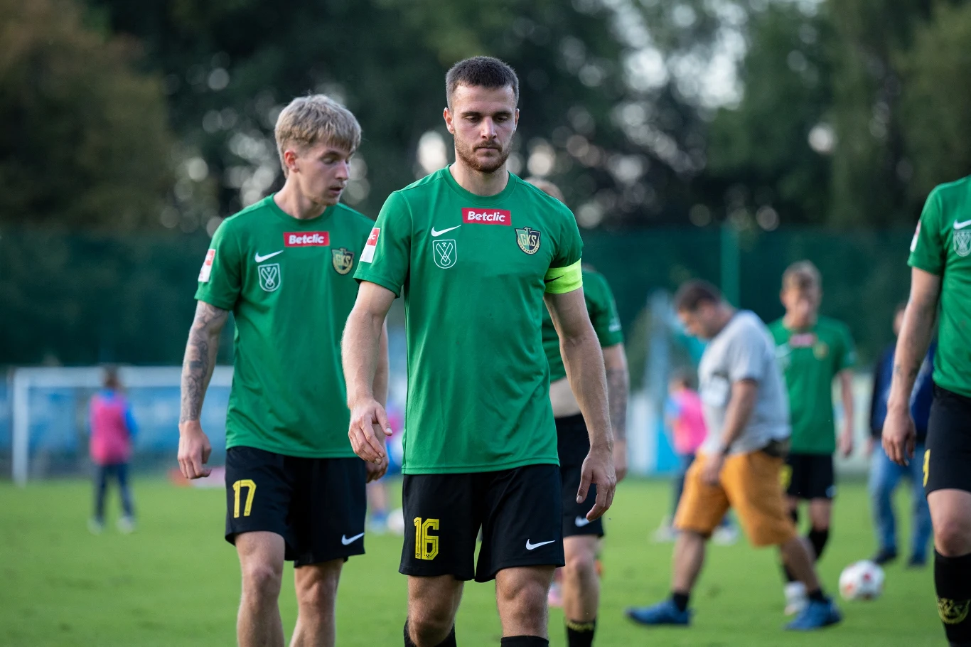 Dwóch piłkarzy w zielonych koszulkach z numerami 17 i 16 idzie po murawie, na pierwszym planie zawodnik z opaską kapitańską, w tle inni gracze oraz rozmyta postać sędziego lub trenera.