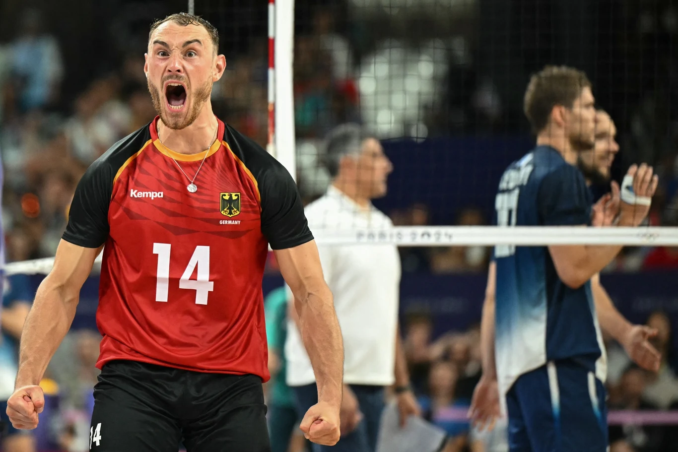 siatkarz w czerwono-czarnym stroju z numerem 14 wyraża silne emocje, stojąc blisko siatki, podczas gdy zawodnicy drużyny przeciwnej w niebieskich strojach są w tle