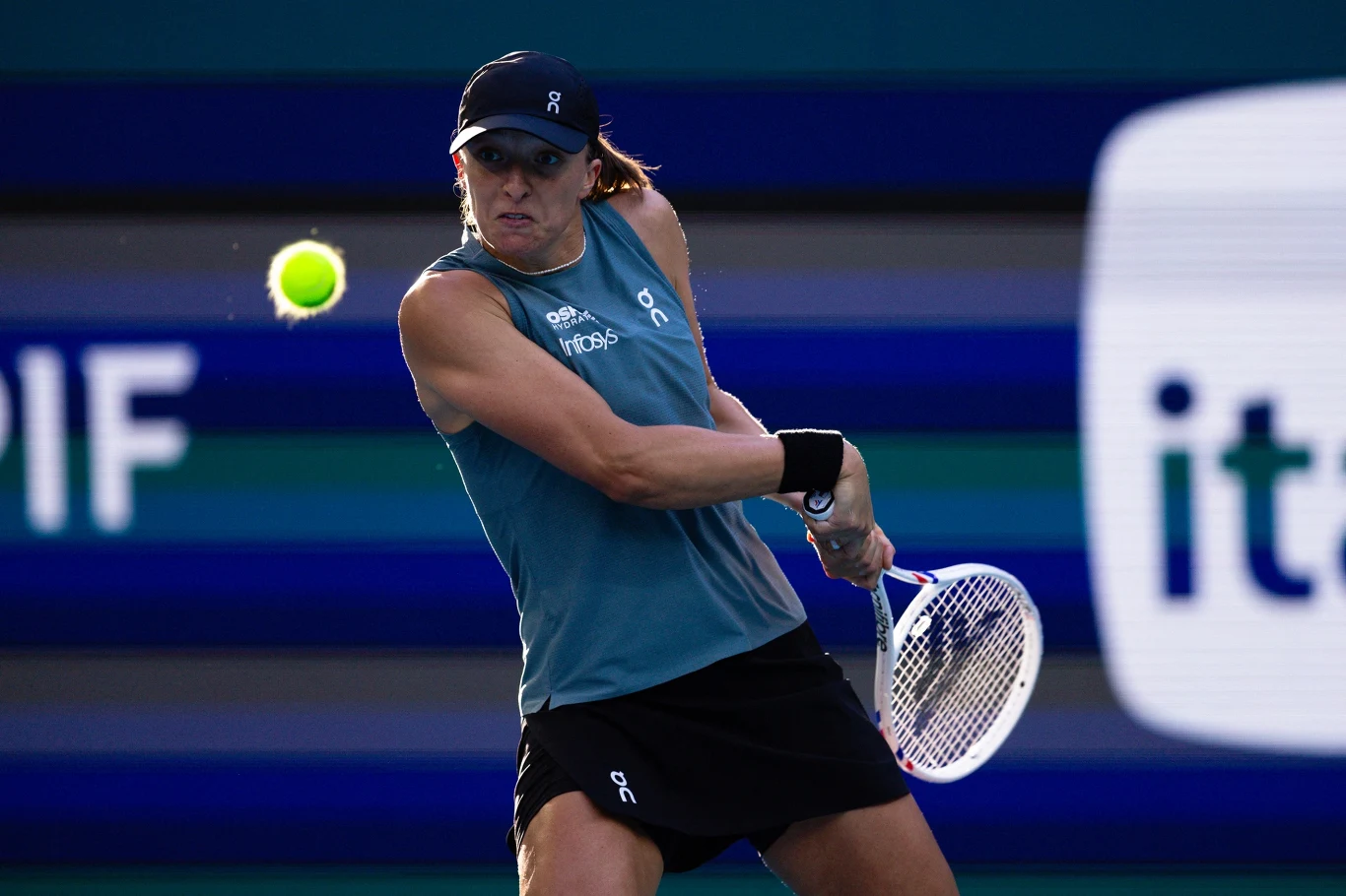 Tenisistka w niebieskiej koszulce i czapce wykonuje dwuręczny bekhend, skupiona na nadlatującej piłce podczas meczu na korcie, z tłem w odcieniach niebieskiego.