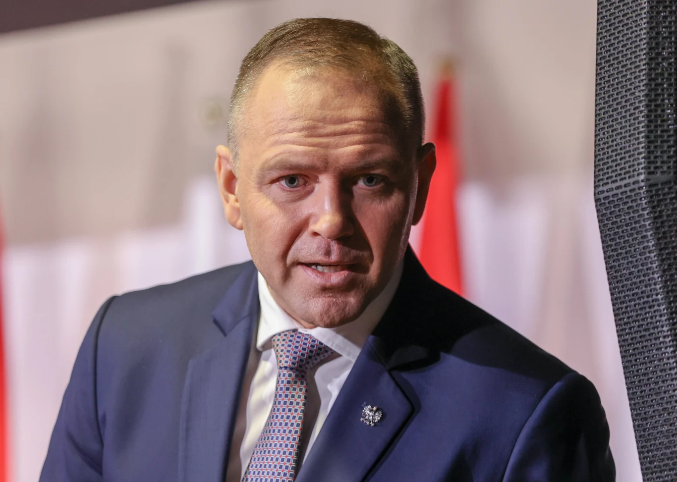 Mężczyzna w granatowym garniturze i krawacie, z krótkimi włosami, stoi na tle biało-czerwonych flag.