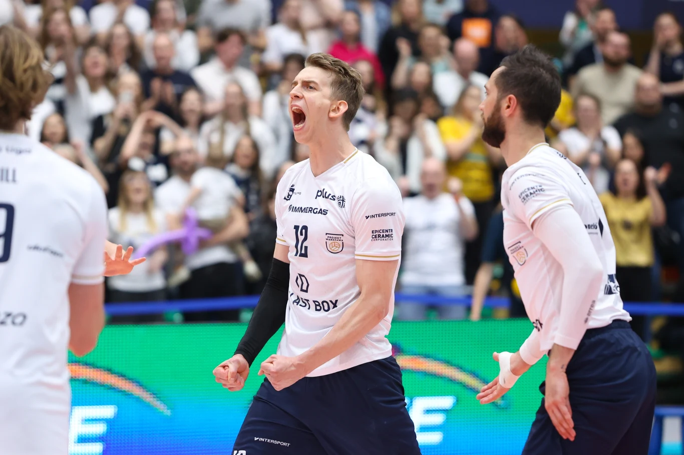 siatkarz w białej koszulce z numerem 12 z okrzykiem radości na twarzy świętuje punkt razem z drużyną, w tle kibice z uniesionymi rękami