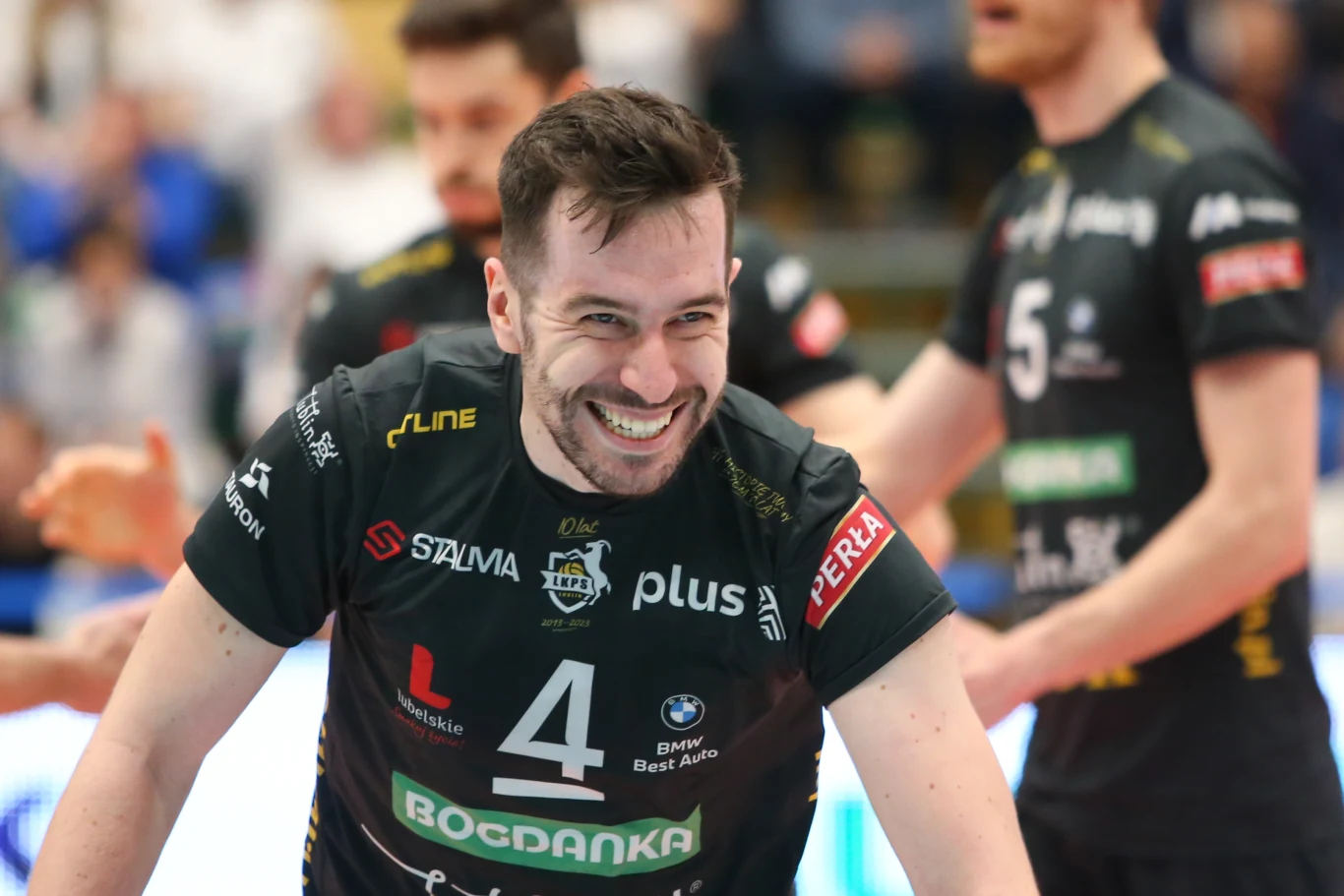 Siatkarz w czarnej koszulce z numerem 4 uśmiecha się szeroko podczas meczu, wokół niego widoczni są inni zawodnicy w podobnych strojach sportowych.