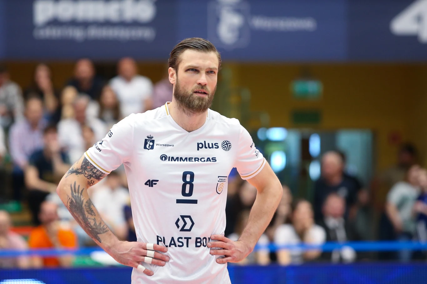 siatkarz w białym stroju z numerem 8 stoi z rękami na biodrach na tle hali sportowej, wokół niego widoczni kibice na trybunach