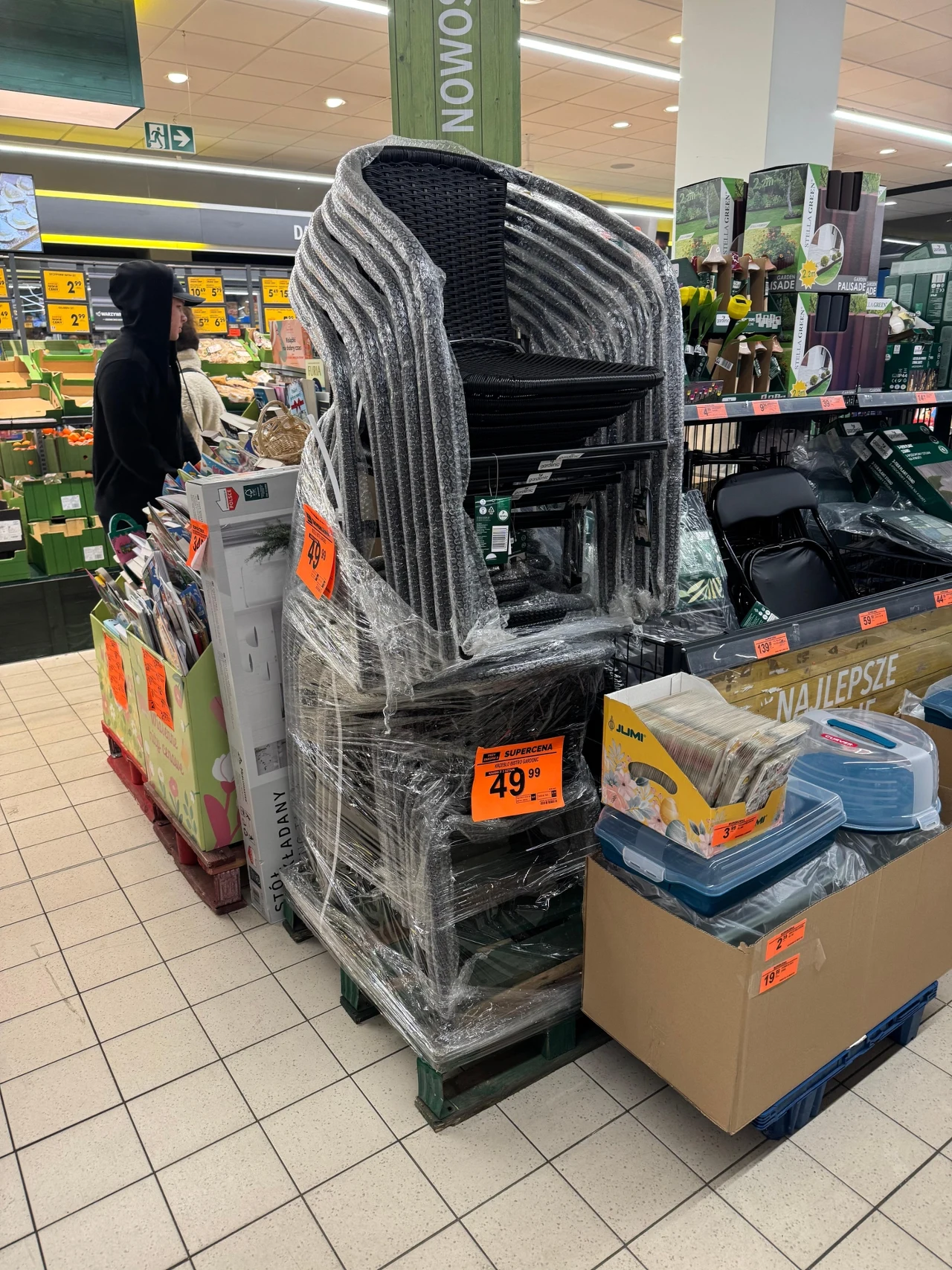 Stos czarnych krzeseł ogrodowych owiniętych folią stretch ustawiony na palecie, otoczony promocjami na artykuły sezonowe w supermarkecie, obok opakowania plastikowych pojemników i ogrodowe akcesoria z widocznymi pomarańczowymi cenówkami.