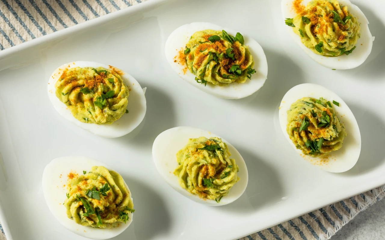 Połówki jajek faszerowane kremowym nadzieniem z awokado, udekorowane świeżymi ziołami i delikatnie posypane przyprawami, ułożone na białym prostokątnym talerzu.