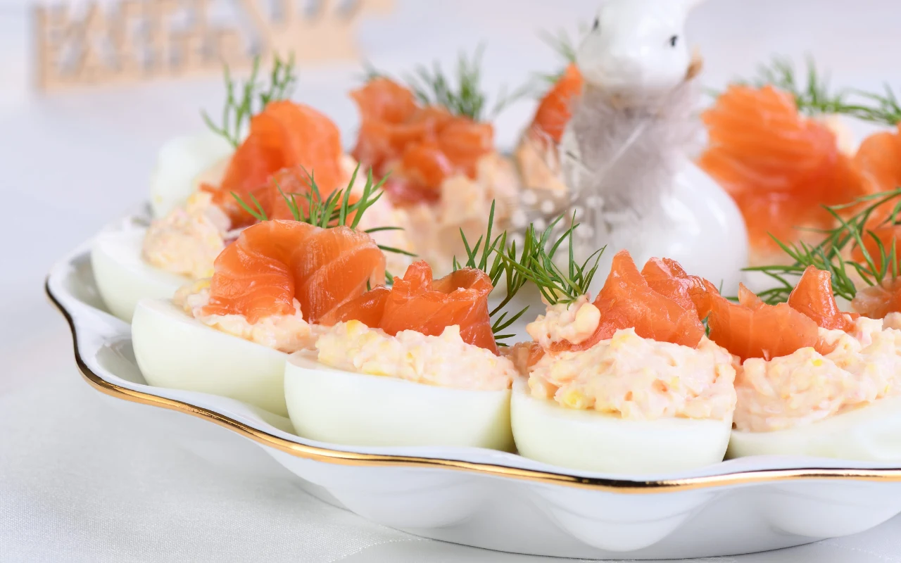 Połówki jajek faszerowane kremowym nadzieniem, udekorowane plastrami łososia i gałązkami koperku, ułożone na eleganckiej białej paterze, w tle ceramiczna dekoracja w kształcie zająca.