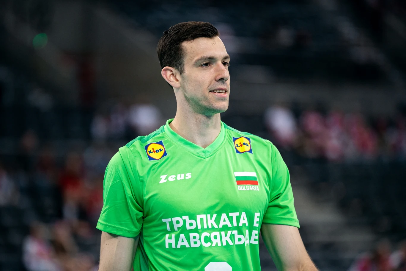 siatkarz w zielonej koszulce z logo sklepu Lidl oraz flagą Bułgarii, stojący na boisku podczas meczu, z rozmytym tłem trybun wypełnionych kibicami