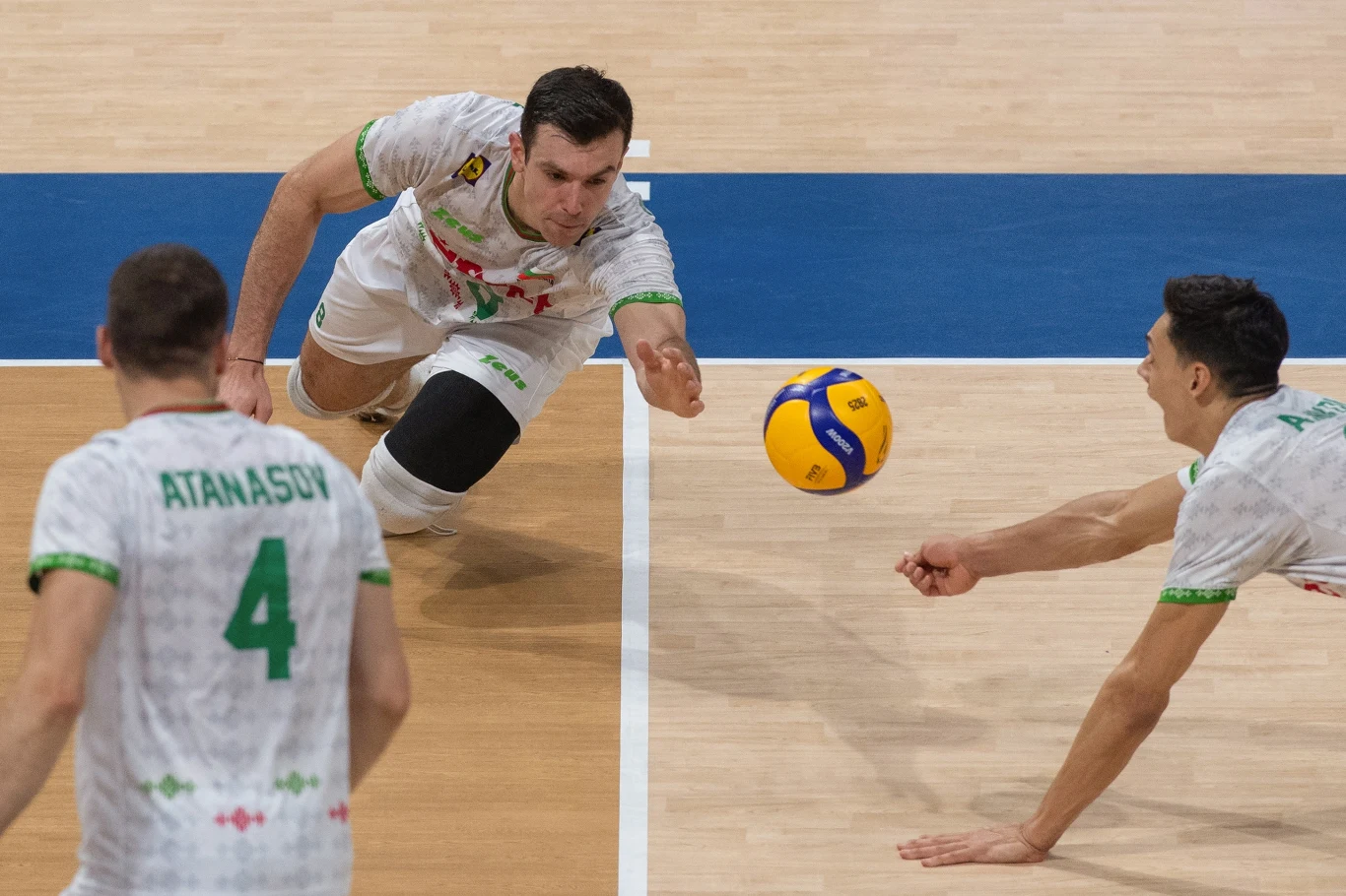 Trzech siatkarzy w białych strojach walczy o piłkę podczas dynamicznej akcji na hali, jeden z zawodników rzuca się w stronę piłki, by ją obronić.