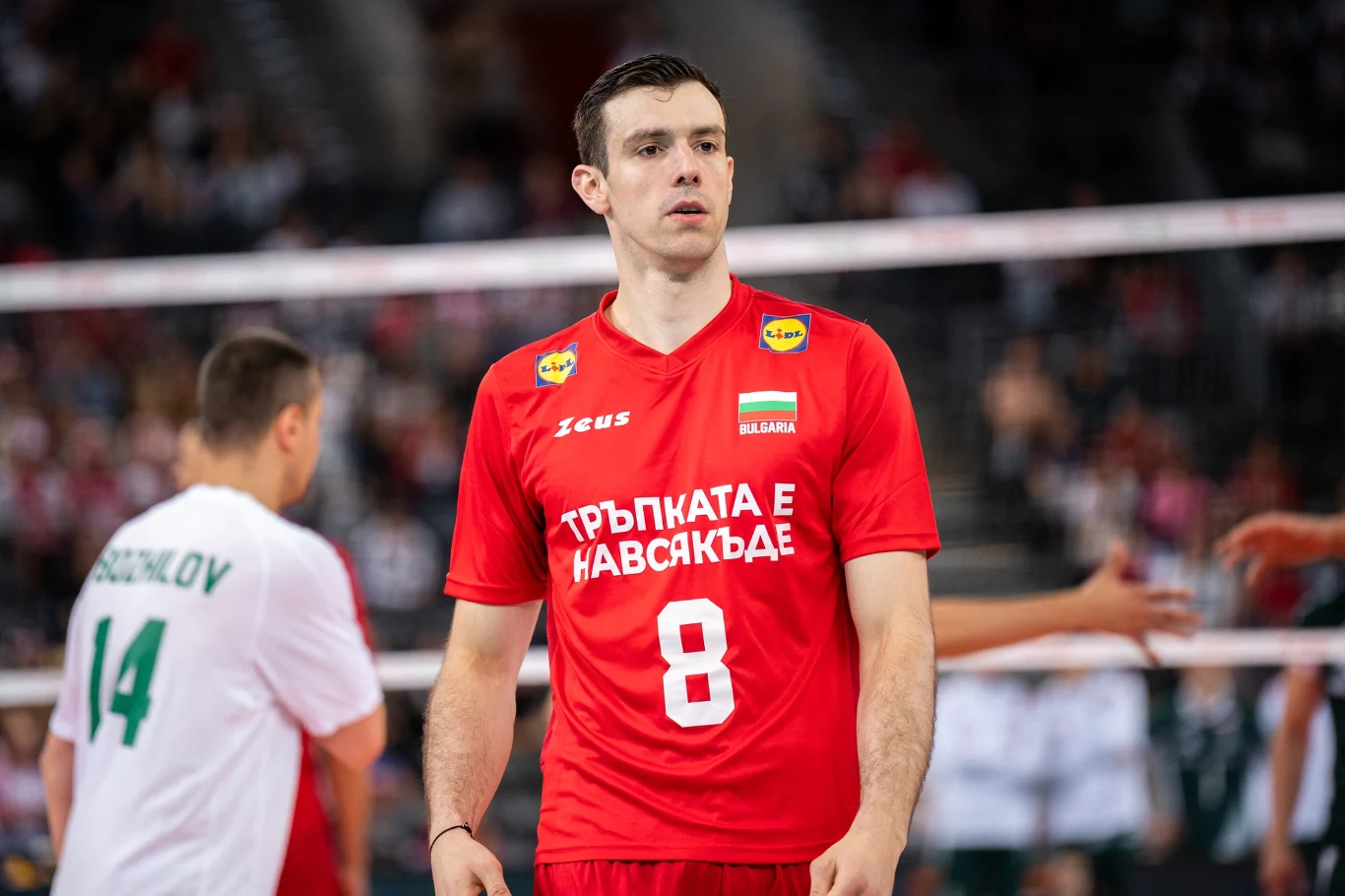siatkarz w czerwonym stroju z numerem 8 i bułgarską flagą na piersi stoi na boisku podczas meczu, w tle inny zawodnik w białej koszulce z numerem 14