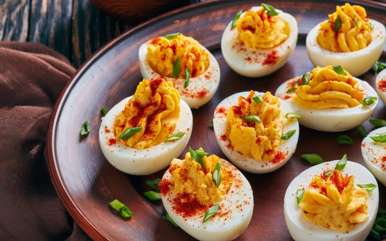 Połówki jajek z kremowym farszem udekorowane posypką z papryki i świeżymi kawałkami szczypiorku, ułożone na brązowym talerzu.