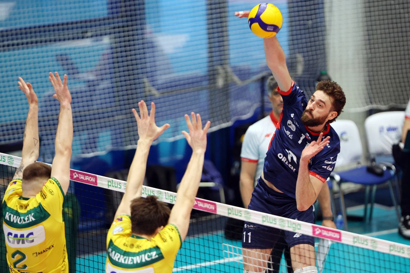 zawodnik w granatowym stroju wykonuje atak siatkarski, uderzając piłkę nad siatką, podczas gdy dwóch przeciwników w żółto-zielonych koszulkach próbuje zablokować akcję wyciągniętymi rękami