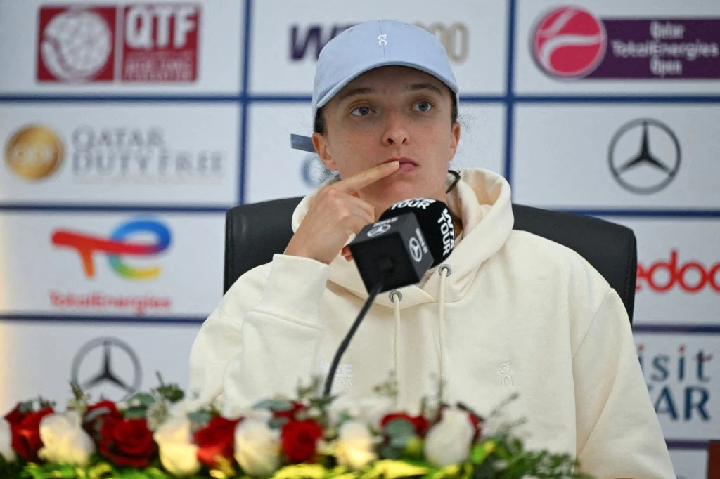 kobieta w jasnej bluzie z kapturem i jasnoniebieskiej czapce z daszkiem siedzi przy stole konferencyjnym z mikrofonem, przed nią leży bukiet czerwono-białych róż, w tle widoczne są logotypy sponsorów turnieju tenisowego