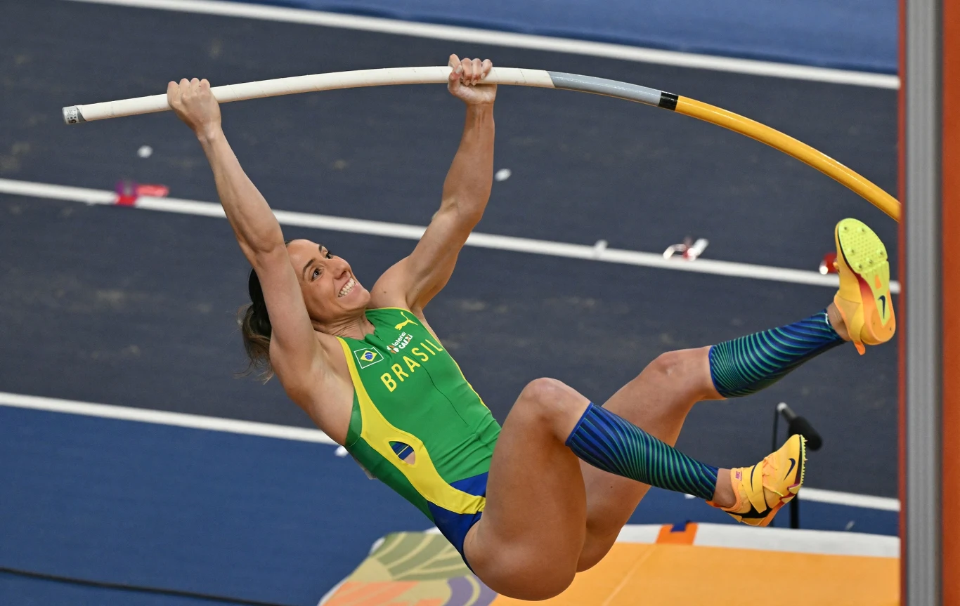 lekkoatletka w zielono-niebieskim stroju z napisem 'Brasil' wykonująca skok o tyczce na stadionie lekkoatletycznym, trzymająca się mocno żółto-białej tyczki w trakcie dynamicznego wybicia nad poprzeczką