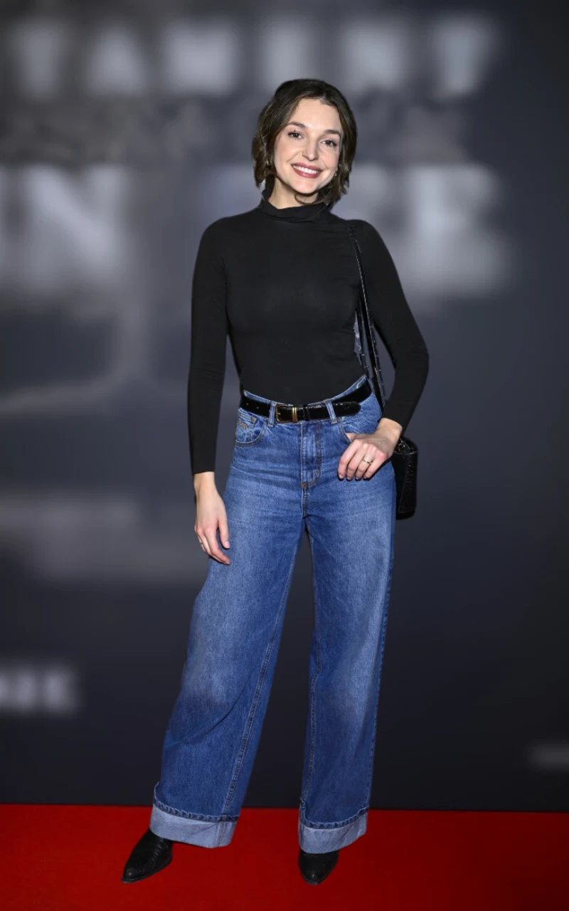 młoda kobieta w czarnym golfie i szerokich niebieskich dżinsach z zamszowym paskiem oraz czarną torebką na ramieniu stoi na czerwonym dywanie, uśmiechnięta, z jedną ręką w kieszeni
