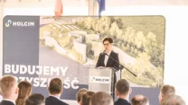 Kobieta w eleganckim stroju przemawia przy mównicy z logo Holcim na tle planszy przedstawiającej inwestycję i hasło „Budujemy przyszłość”; w pierwszym planie zebrani słuchacze w garniturach.