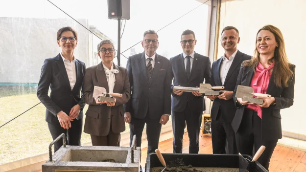 Sześć osób w eleganckich strojach stoi obok siebie uśmiechając się, trzymając w rękach kielnie z zaprawą, przy dwóch pojemnikach z betonem oznaczonych napisem 'Cementownia Kujawy', co sugeruje uroczystą inaugurację lub ważne wydarzenie związane z budow...