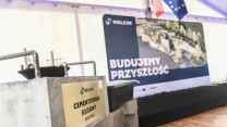 Kamień węgielny z tabliczką 'Cementownia Kujawy', ustawiony na betonowej konstrukcji, na dalszym planie scena z dużą planszą z napisem 'Budujemy przyszłość', obok której widoczne są flagi Polski i Unii Europejskiej.