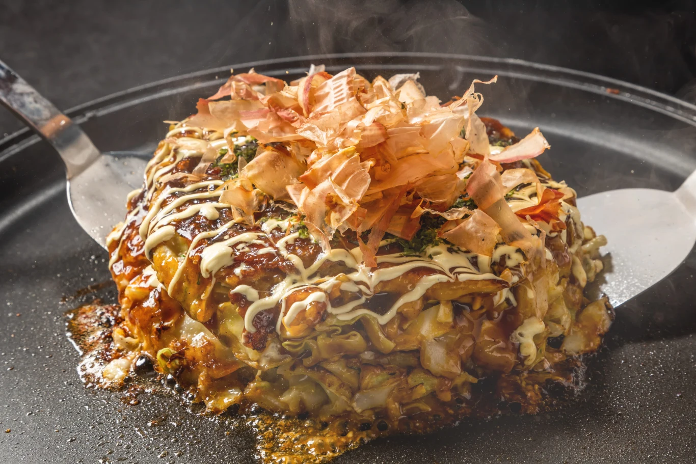 Japoński placek okonomiyaki z kapustą i dodatkami, polany majonezem oraz sosem, udekorowany płatkami suszonego bonito, smażony na blacie teppan, obok dwie łopatki kuchenne.