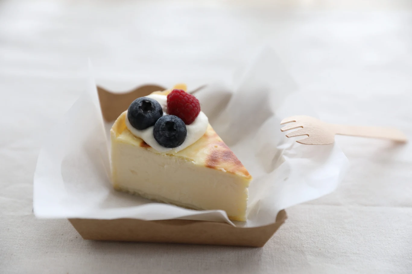 kawałek sernika podany na papierze w tekturowym pojemniku, udekorowany bitą śmietaną, dwoma jagodami i jedną maliną, obok znajduje się drewniany widelec