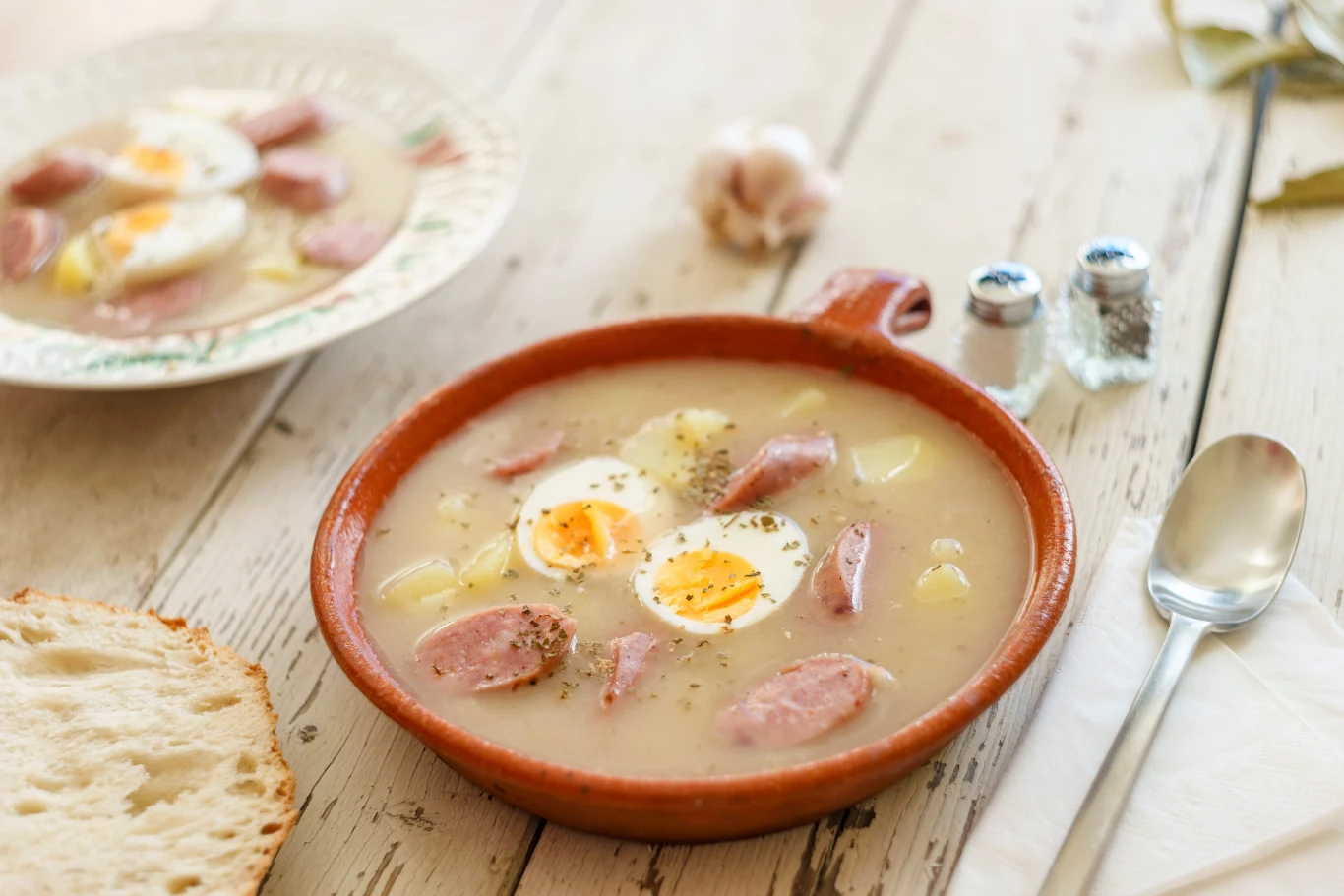 Talerz żurku z kawałkami białej kiełbasy, jajkiem na twardo pokrojonym w plastry oraz ziemniakami, podany na jasnym, drewnianym stole obok kromki chleba, łyżki i przypraw w solniczce i pieprzniczce.
