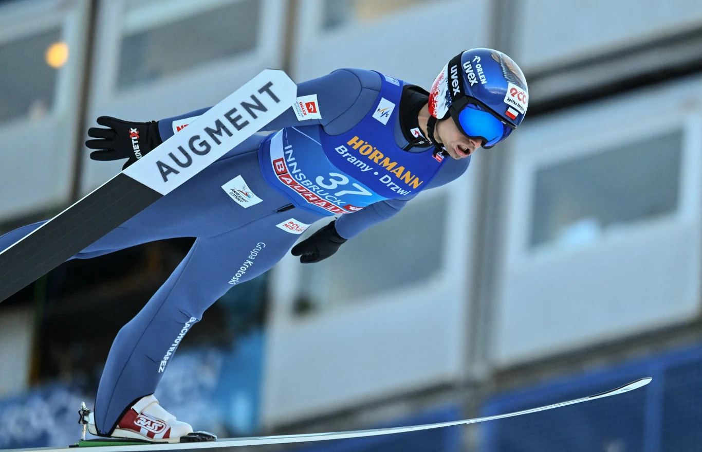 skoczek narciarski w niebieskim kombinezonie leci w powietrzu w trakcie skoku, trzymając ciało w aerodynamicznej pozycji, z profesjonalnym kaskiem i goglami, na tle rozmytych trybun stadionu