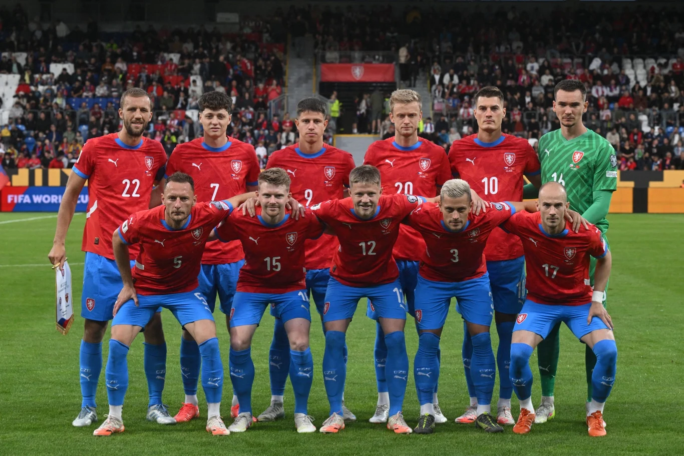 jedenastka piłkarzy w czerwonych koszulkach i niebieskich spodenkach, ustawionych w dwóch rzędach na boisku piłkarskim przed rozpoczęciem meczu, na tle zapełnionych trybun