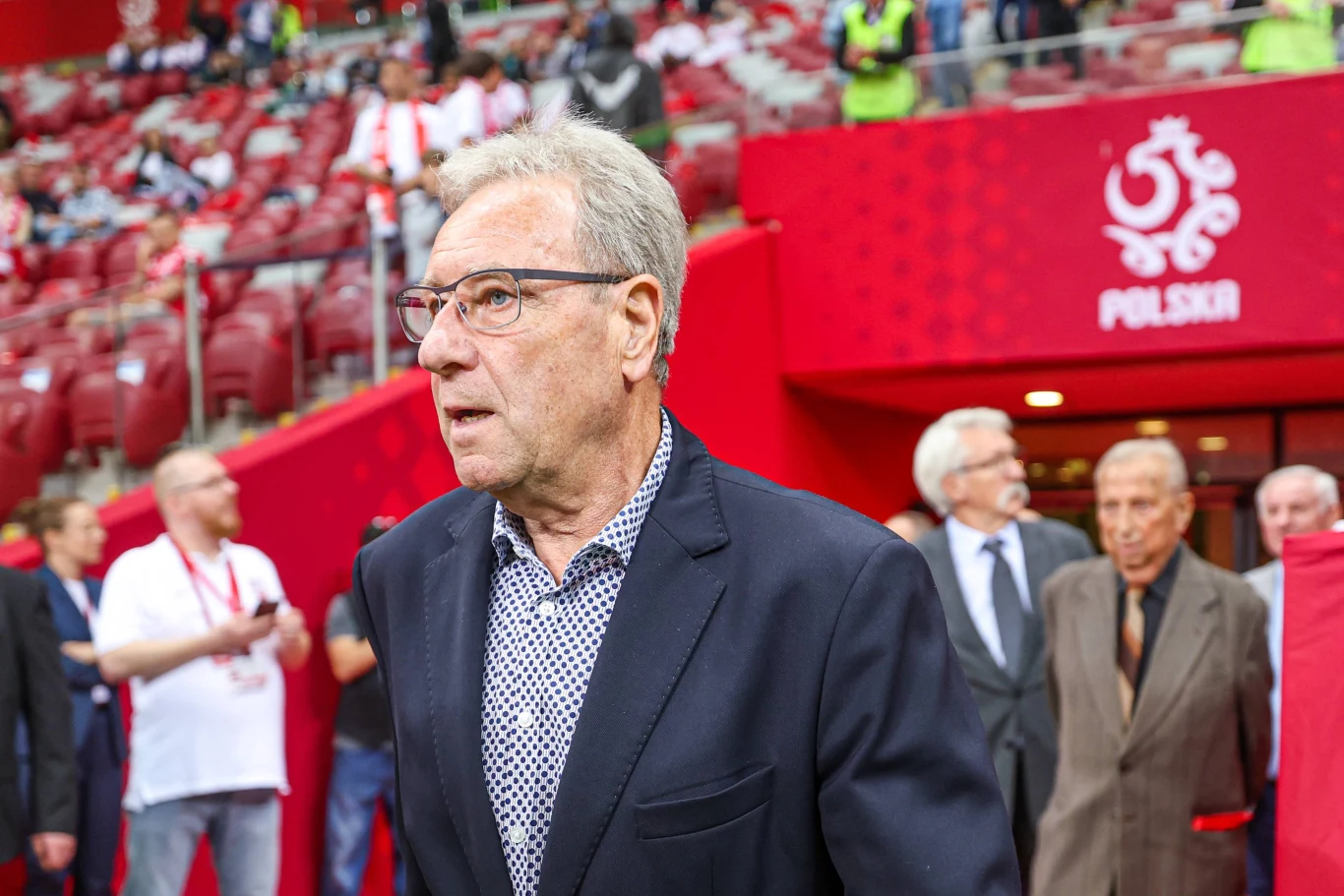 starszy mężczyzna w garniturze idzie wzdłuż trybun stadionu piłkarskiego, w tle widoczne są osoby w eleganckich strojach oraz czerwona strefa z napisem „Polska” i godłem narodowym