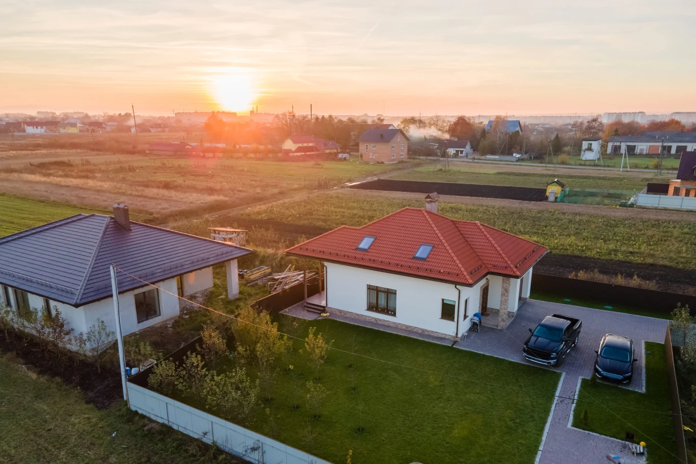 Parterowy dom z czerwonym dachem otoczony zadbanym ogrodem, dwa zaparkowane samochody na podjeździe, obok drugi dom z ciemniejszym dachem, w tle rozległe pola i zachodzące słońce.