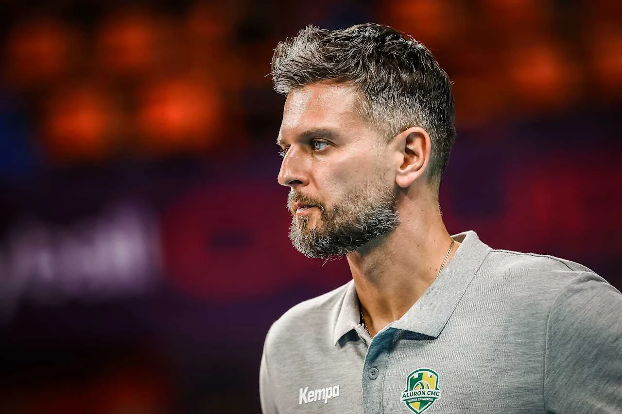 Mężczyzna z krótkimi, siwiejącymi włosami i brodą, ubrany w szarą koszulkę polo z logo drużyny sportowej oraz napisem 'Kempa', znajduje się na kolorowo rozmazanym tle, patrząc w bok z poważnym wyrazem twarzy.