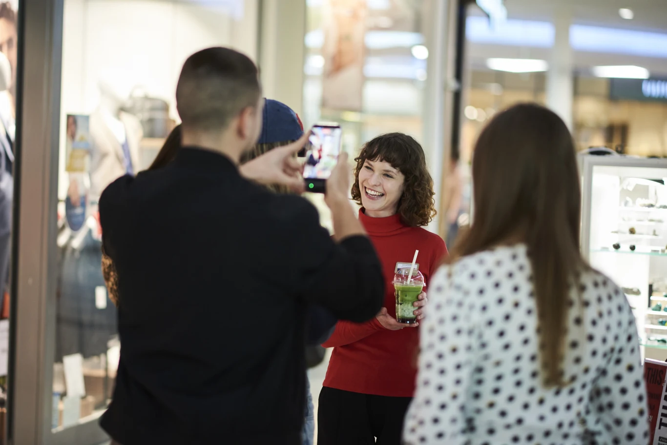 Grupa młodych osób spędza czas w galerii handlowej; kobieta w czerwonym swetrze z napojem w ręce uśmiecha się, podczas gdy jedna z osób robi jej zdjęcie smartfonem.