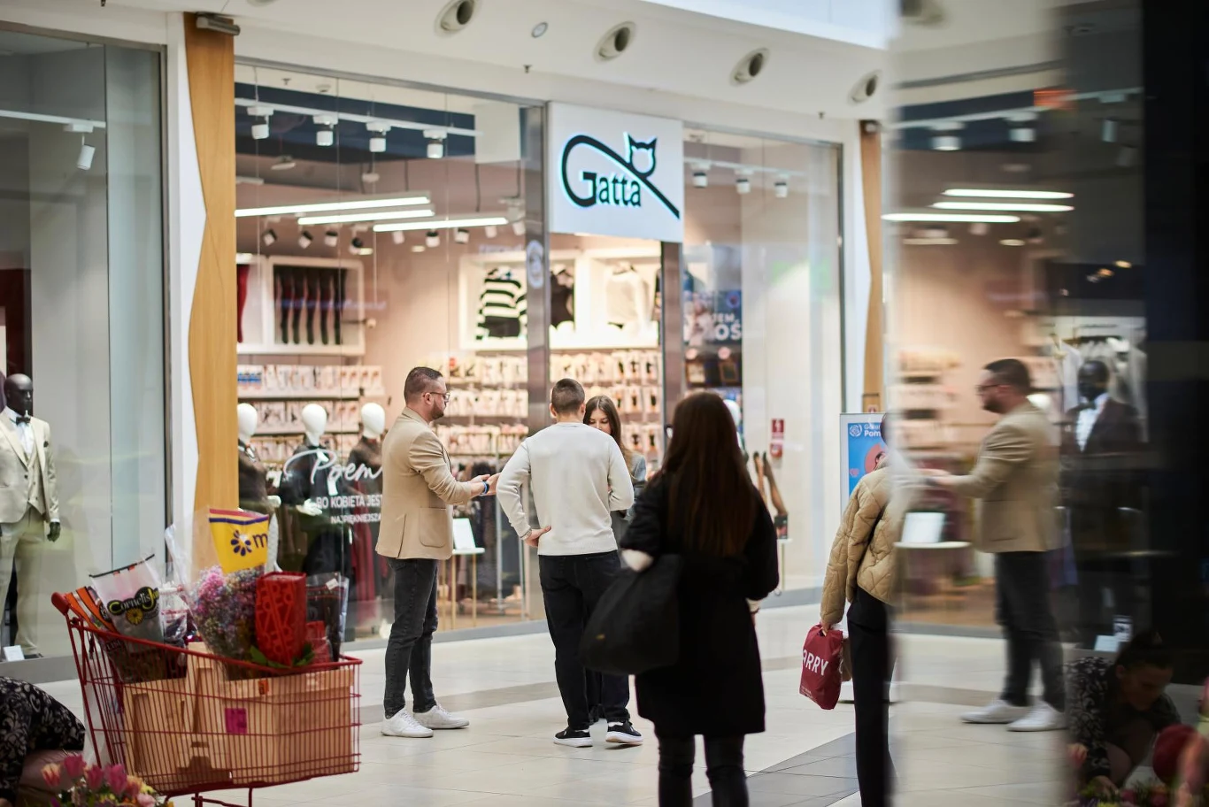 grupa osób stoi przed sklepem Gatta w centrum handlowym, w tle witryna z bielizną i manekinami, na pierwszym planie przechodzący klienci oraz wózek z zakupami
