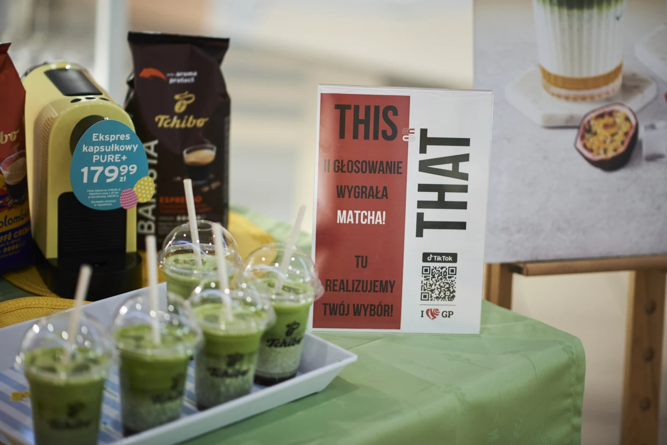 na zielonym obrusie stoi tacka z pięcioma plastikowymi kubkami z napojem matcha z lodem, obok ekspres do kawy Tchibo, opakowania kawy oraz tablica z napisem THIS THAT i informacją o wygranej matchy w głosowaniu