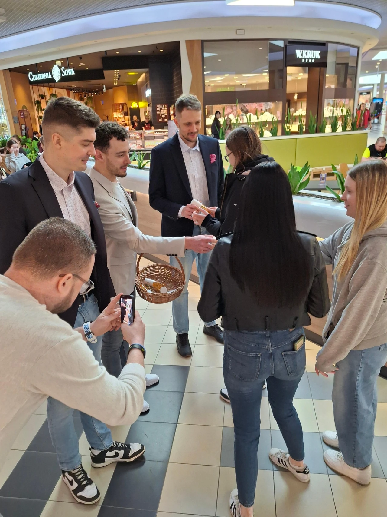 Grupa młodych osób w centrum handlowym, mężczyzna w marynarce trzyma koszyk i wręcza mały przedmiot jednej z kobiet, inni obserwują i robią zdjęcia smartfonami.