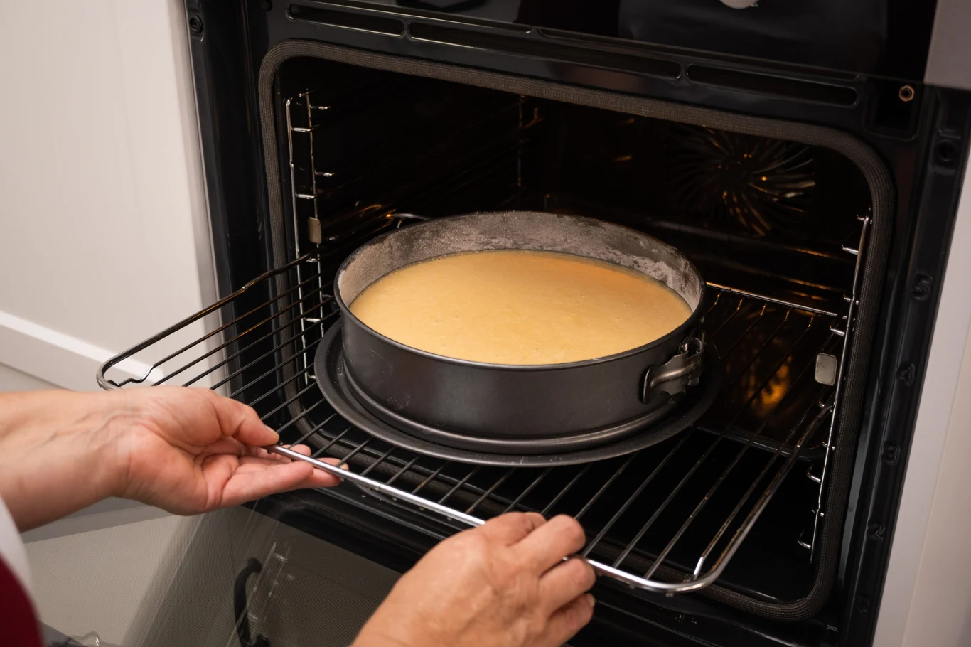 Surowa masa sernika w okrągłej formie wstawiana do nagrzanego piekarnika przez osobę trzymającą kratkę ovenową.