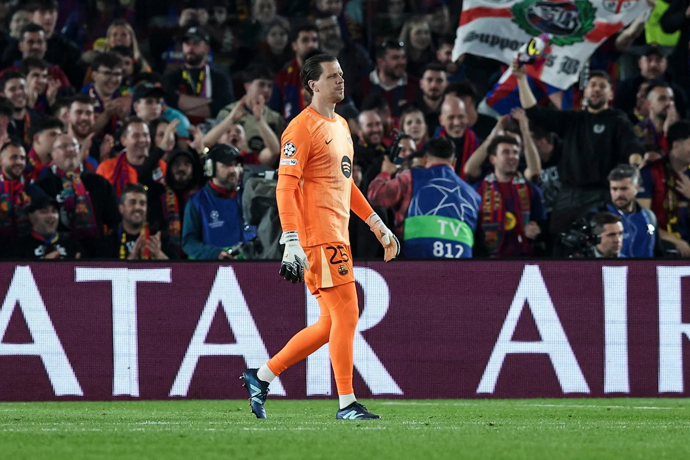 bramkarz w pomarańczowym stroju idący po murawie boiska podczas meczu piłkarskiego, w tle tłum kibiców z flagami i banerami