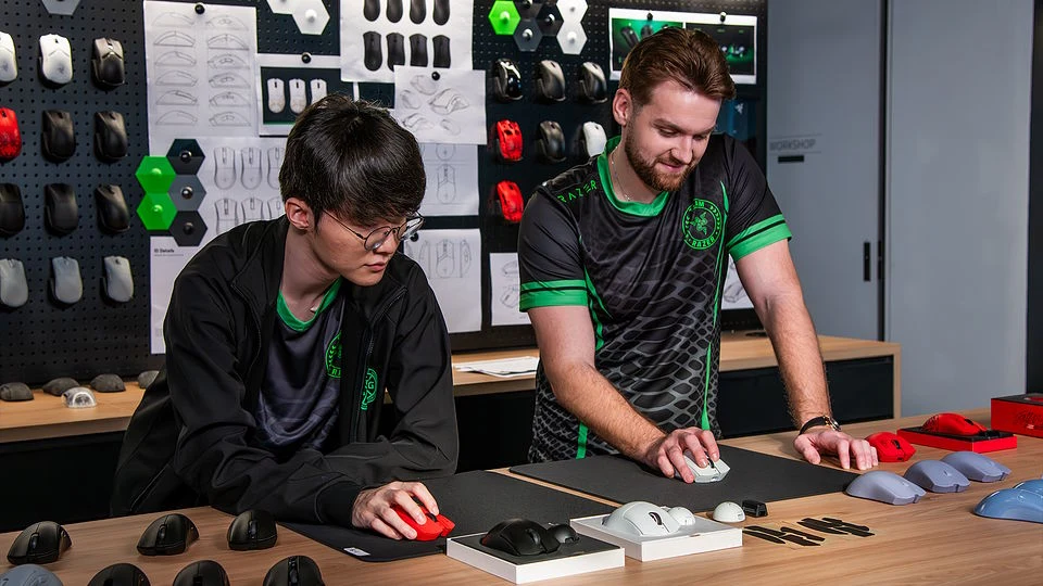 Dwóch mężczyzn testujących różne modele komputerowych myszy na blacie, wokół nich liczne prototypy i projekty myszy umieszczone na ścianie w tle, na obu widoczne sportowe koszulki z zielonymi elementami.