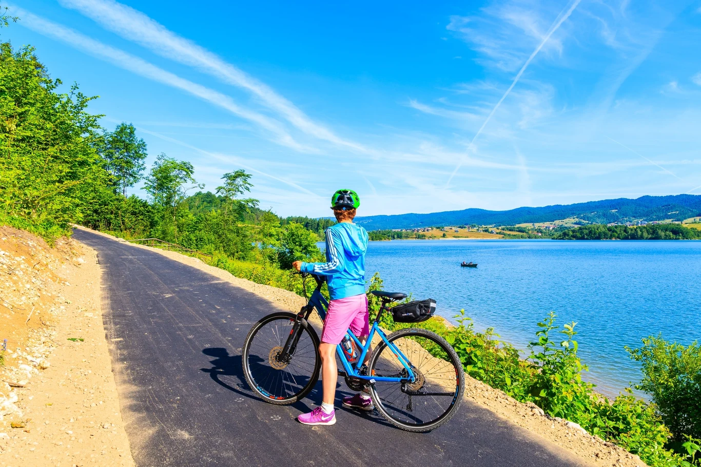 Osoba w kasku rowerowym i sportowym stroju stoi z rowerem przy asfaltowej ścieżce nad dużym jeziorem, otoczona zielenią, z widokiem na góry i błękitne niebo z wyraźnymi smugami chmur.