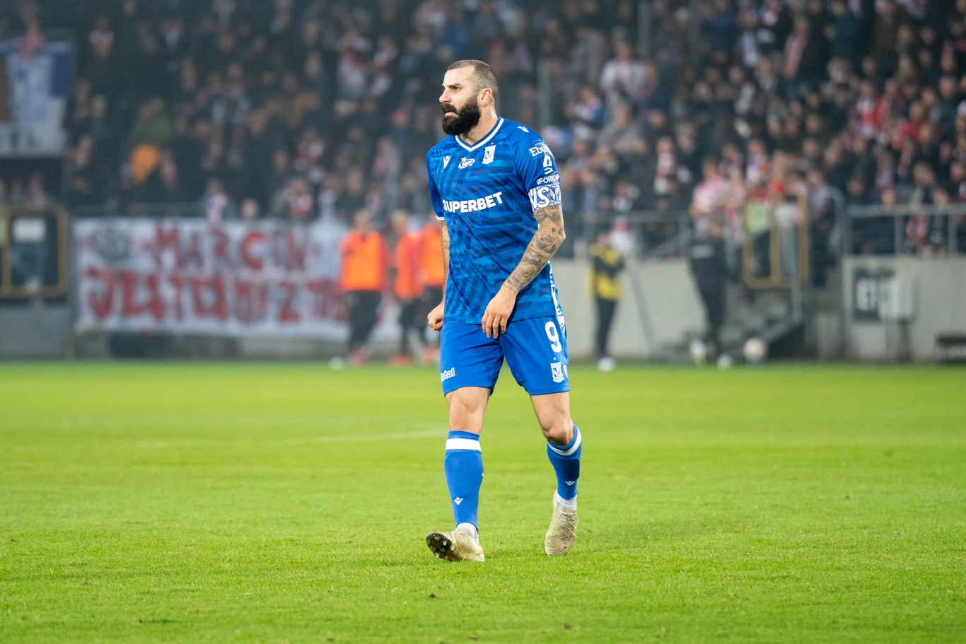 piłkarz w niebieskim stroju z widocznymi tatuażami na rękach spaceruje po murawie stadionu, w tle rozmyty tłum kibiców i transparent