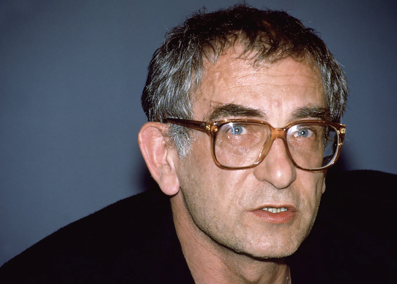 mężczyzna w średnim wieku z krótkimi, siwiejącymi włosami i dużymi okularami o grubych oprawkach patrzy przed siebie na neutralnym tle