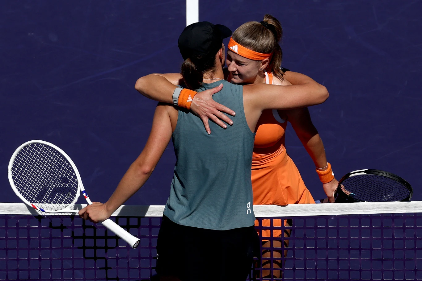 Dwie tenisistki obejmują się przy siatce, trzymając rakiety w dłoniach, obie ubrane w sportowe stroje, jedna w pomarańczowym zestawie i opasce, druga w czarnej czapce i oliwkowym topie, wyrażając sportową solidarność po zakończonym meczu.