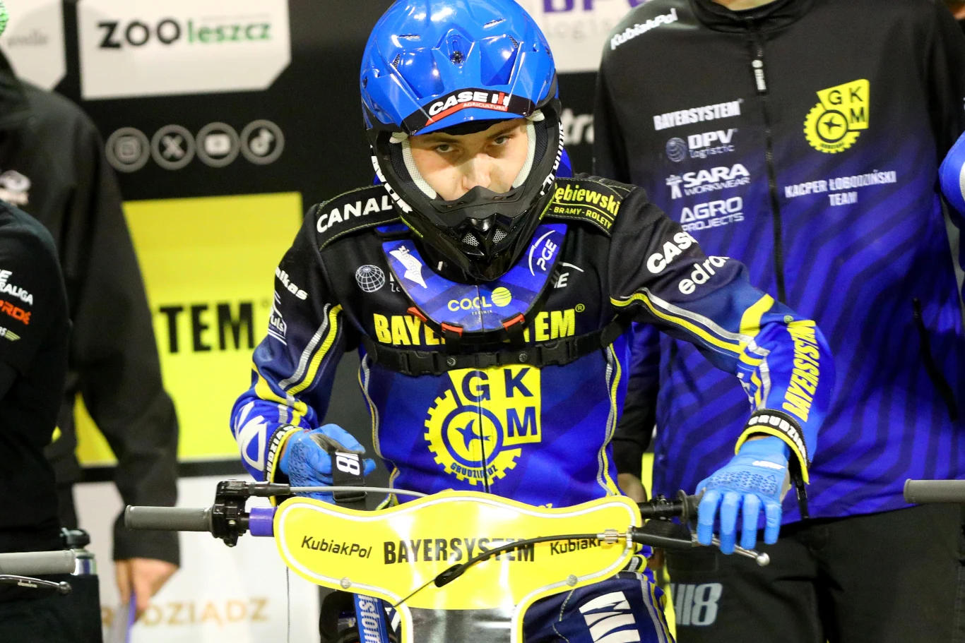 zawodnik żużlowy w niebieskim kasku i kombinezonie z logo GKM Grudziądz stoi przy motocyklu na tle drużynowych barw i sponsorów
