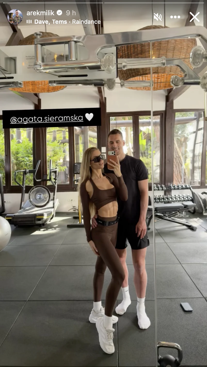 kobieta w sportowym, brązowym stroju i mężczyzna w czarnej koszulce oraz szortach pozują razem przed lustrem na siłowni; w tle nowoczesny sprzęt do ćwiczeń i duże okna wpuszczające naturalne światło