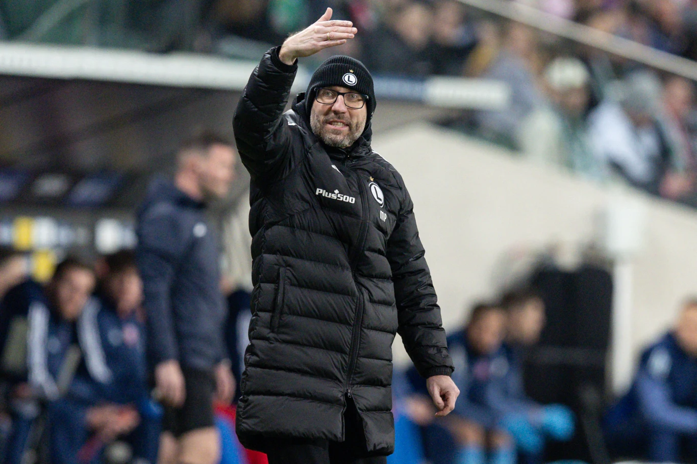 Marek Papszun Trener w czarnej kurtce i czapce z logo klubu piłkarskiego gestykuluje ręką na linii bocznej boiska podczas meczu, w tle widoczni są zawodnicy i sztab szkoleniowy rezerwowi siedzący na ławce.