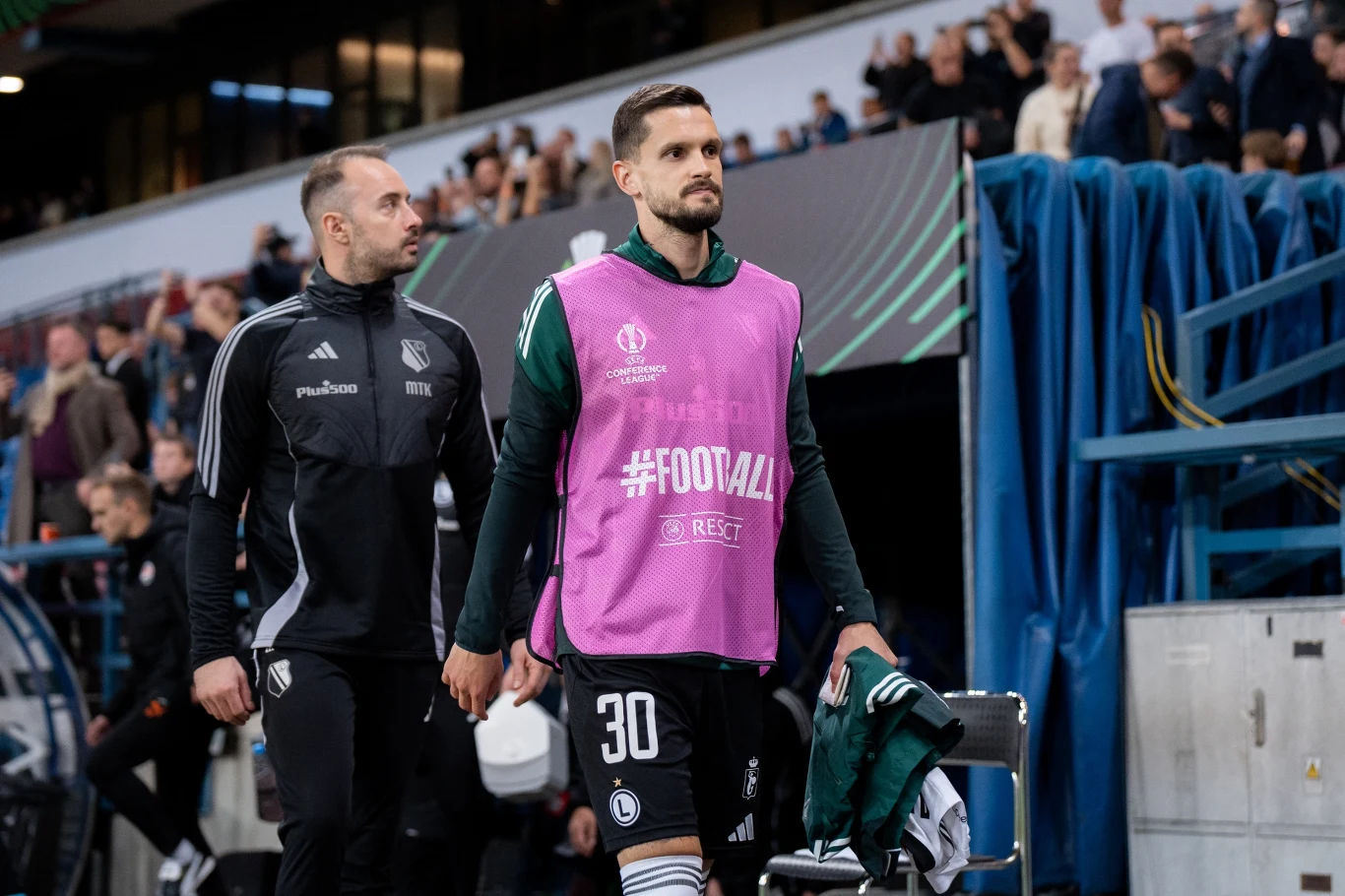 Petar Stojanović piłkarz w zielonej koszulce z numerem 30 i różową kamizelką treningową z napisem #FOOTBALL oraz członek sztabu szkoleniowego w czarnej odzieży klubowej idą obok siebie wzdłuż linii bocznej boiska, w tle trybuny pełne kibiców